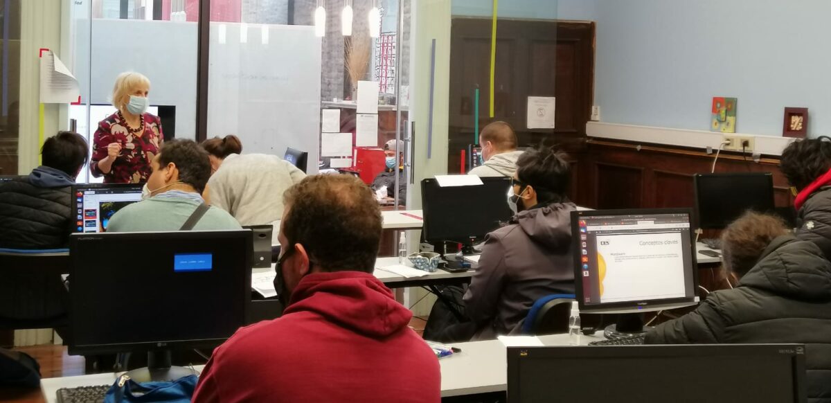 Jóvenes sentados en largas mesas con computadores escuchando a una docente hablar en un salón de clases