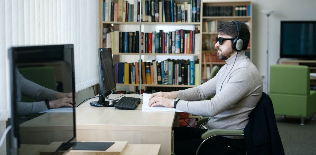 Persona ciega sentada en oficina, usa lentes oscuros y auriculares y lee.