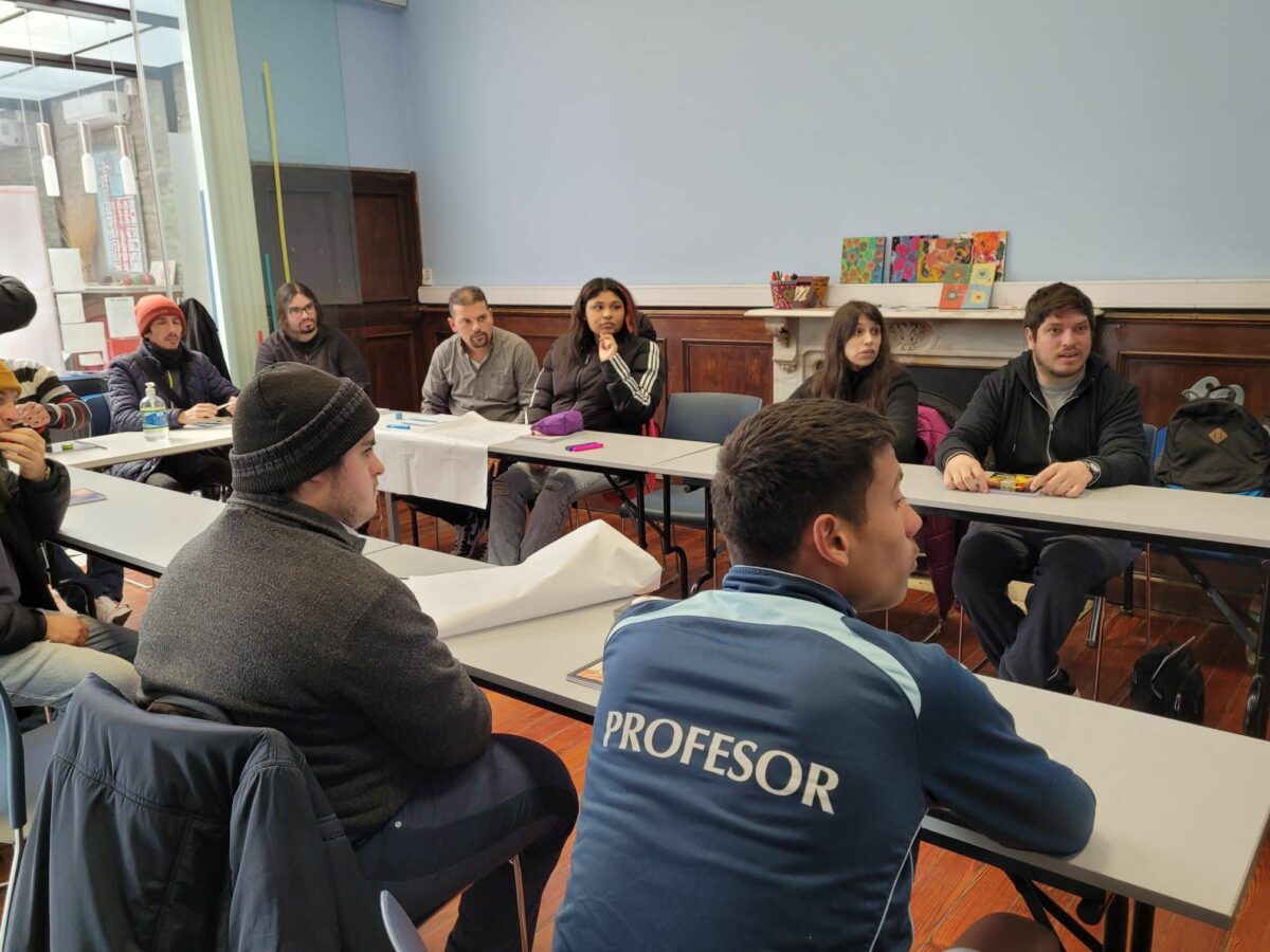 Grupo de estudiantes sentados en mesas en formato "U" en el interior de un salón de clases.
